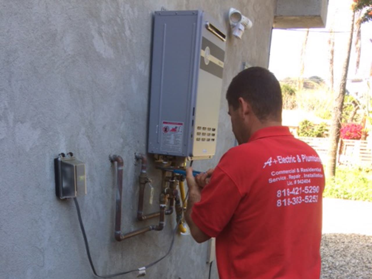 A+ Electric & Plumbing technician in company uniform servicing a tankless water heater at a Los Angeles home.